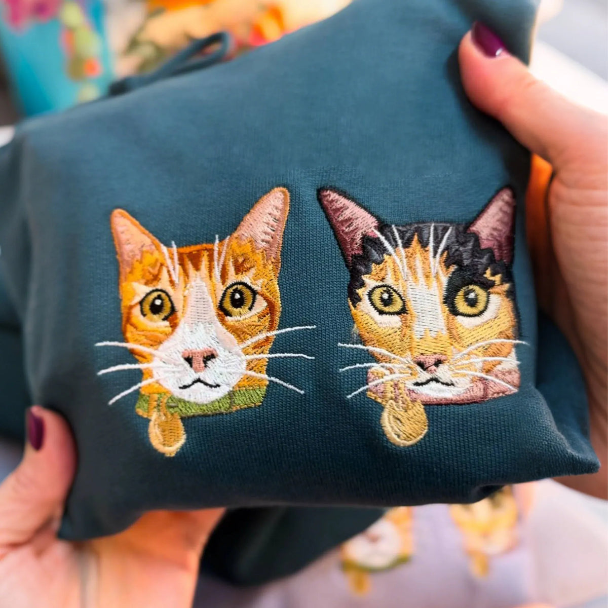 Two orange and calico cat faces embroidered side by side on a teal sweatshirt