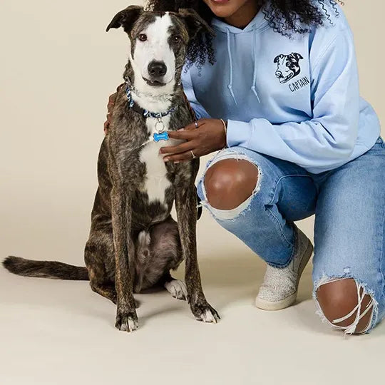 Woman kneeling with her brindle dog, wearing a light blue hoodie with the dog's line art portrait