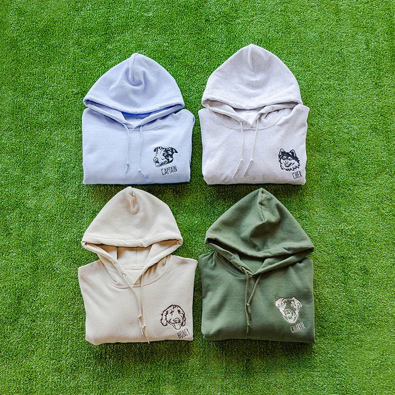 Four pet line art hoodies in lavender, white, sand and green laid out on grass