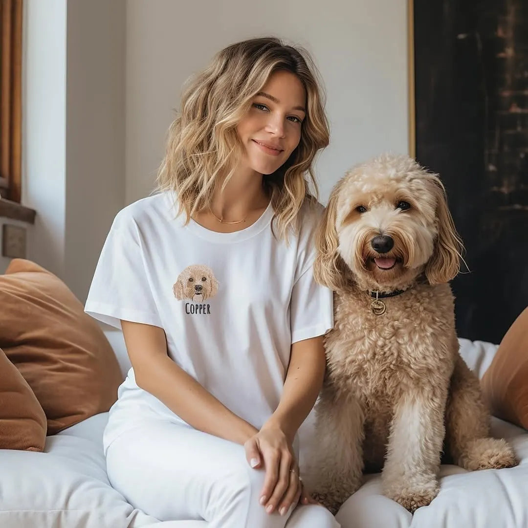 Woman sitting with her goldendoodle, wearing a white tee with a small embroidered doodle portrait