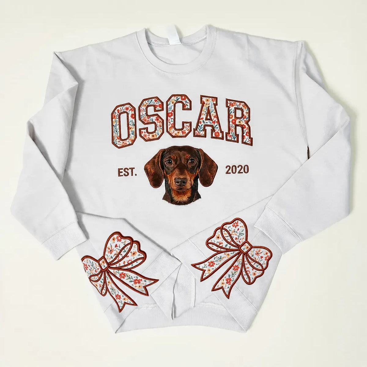 White sweatshirt with OSCAR in red floral letters above an embroidered dachshund and red floral bows on the cuffs