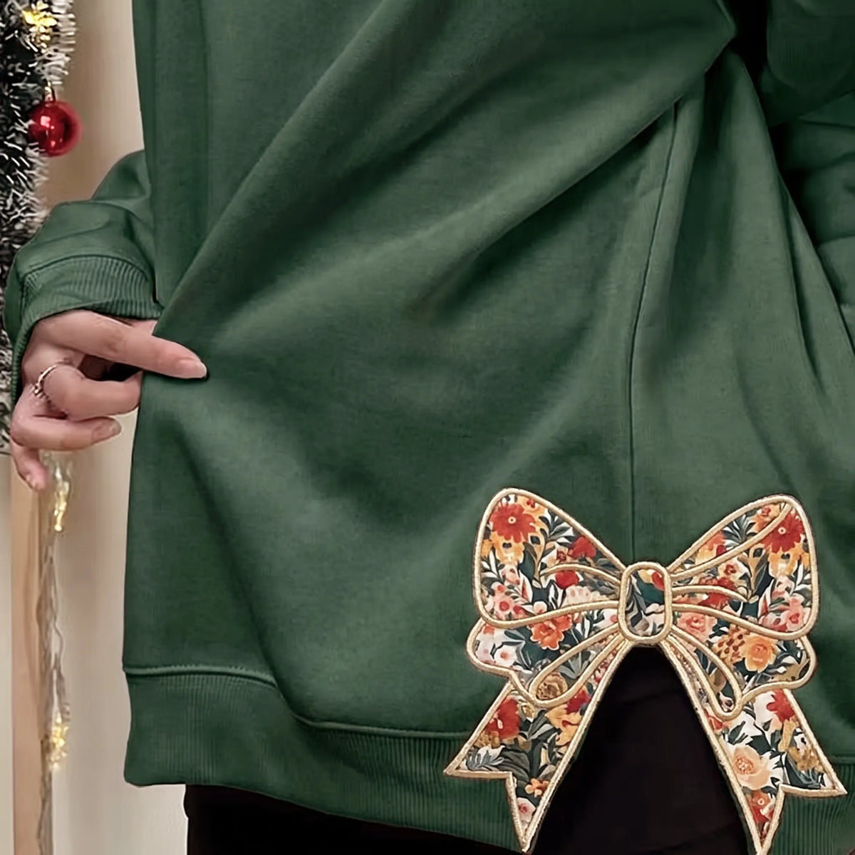 Woman wearing a forest green sweatshirt with a large floral bow appliqué at the hem