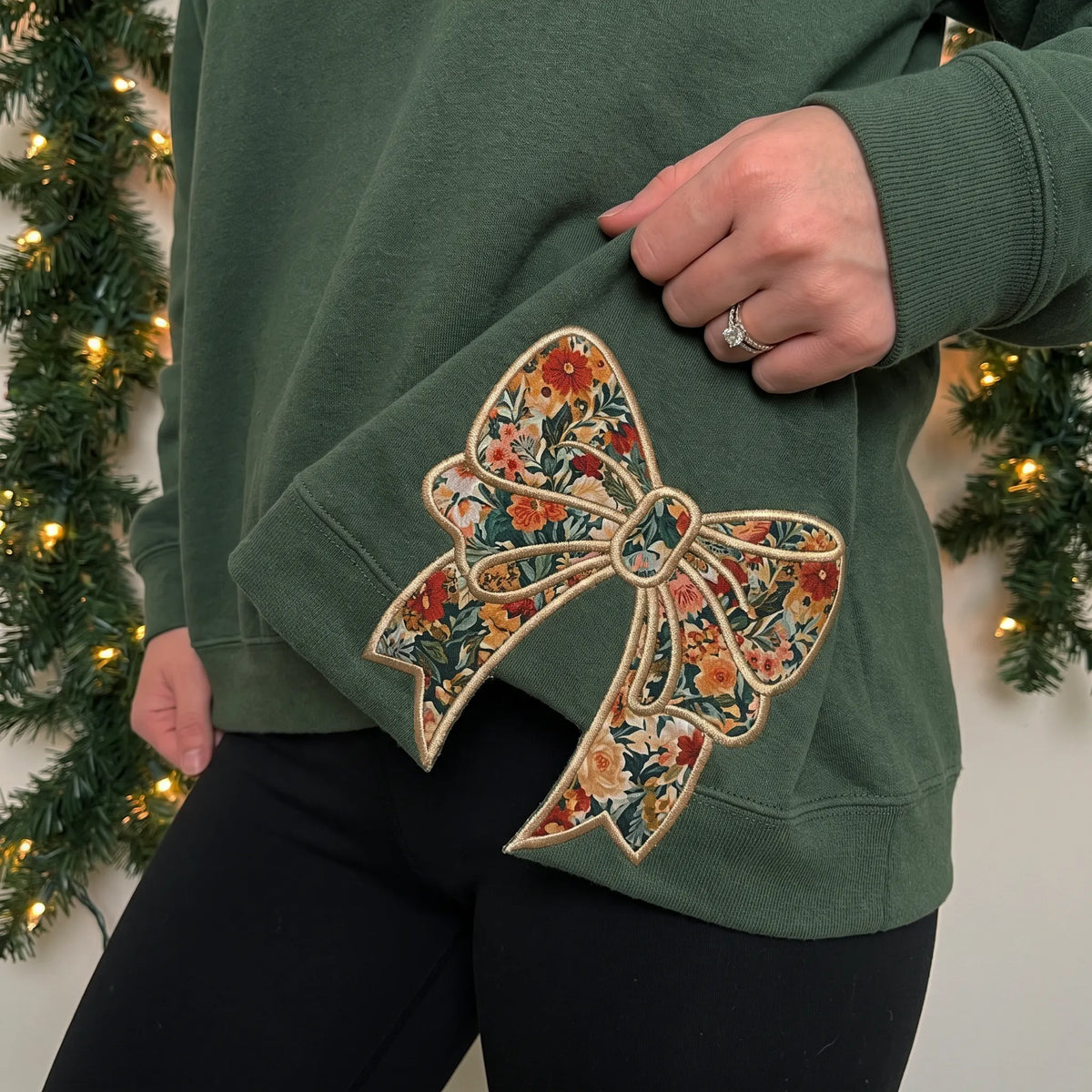 Floral bow appliqué at the hem of a forest green sweatshirt, worn by a woman