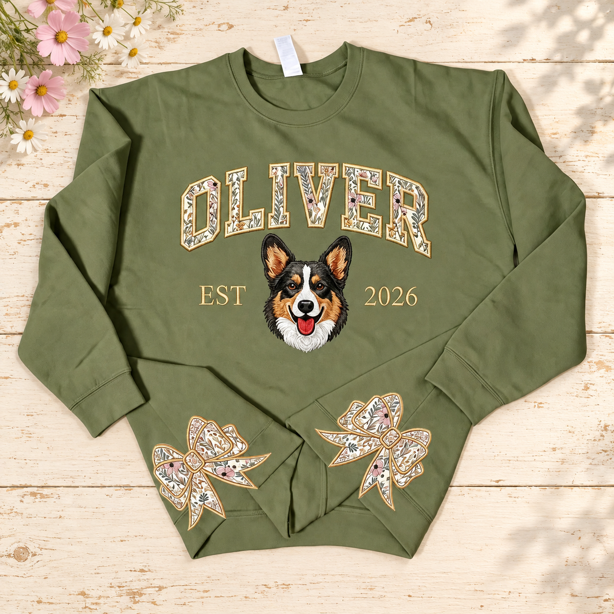 Olive OLIVER pet portrait sweatshirt with floral bow appliques, EST 2026, on a cream garden table with pink flowers