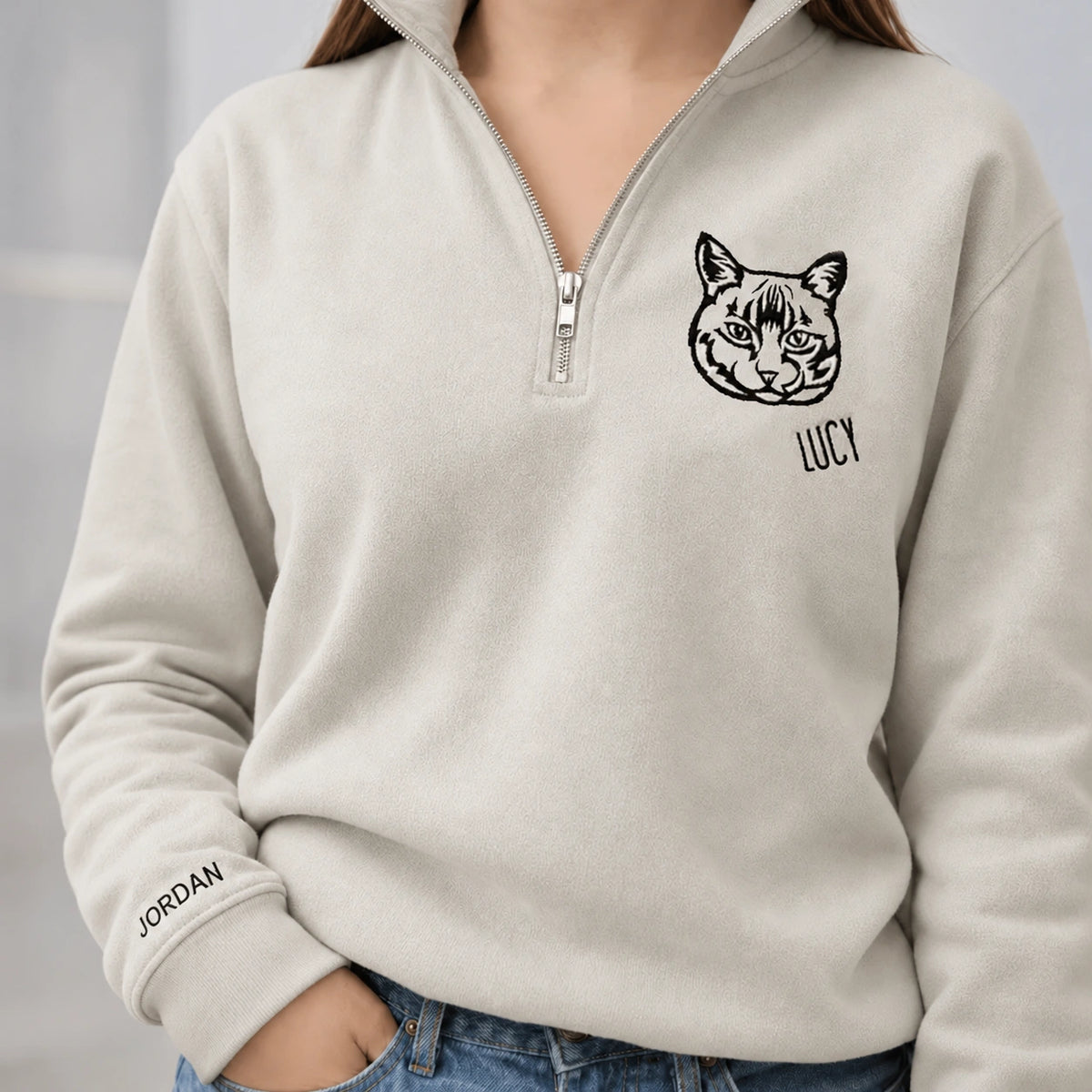 Woman in a light gray quarter zip sweatshirt with a line art cat named LUCY and JORDAN on the cuff